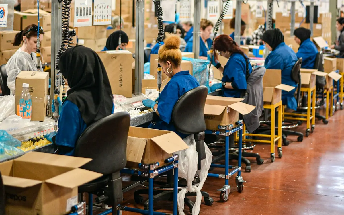 Fotografia de uma linha de montagem com vários trabalhadores sentados em estações de trabalho dispostas em filas. Eles vestem uniformes azuis e alguns usam lenços na cabeça ou toucas de proteção, enquanto caixas e carrinhos de apoio completam o cenário da fábrica.