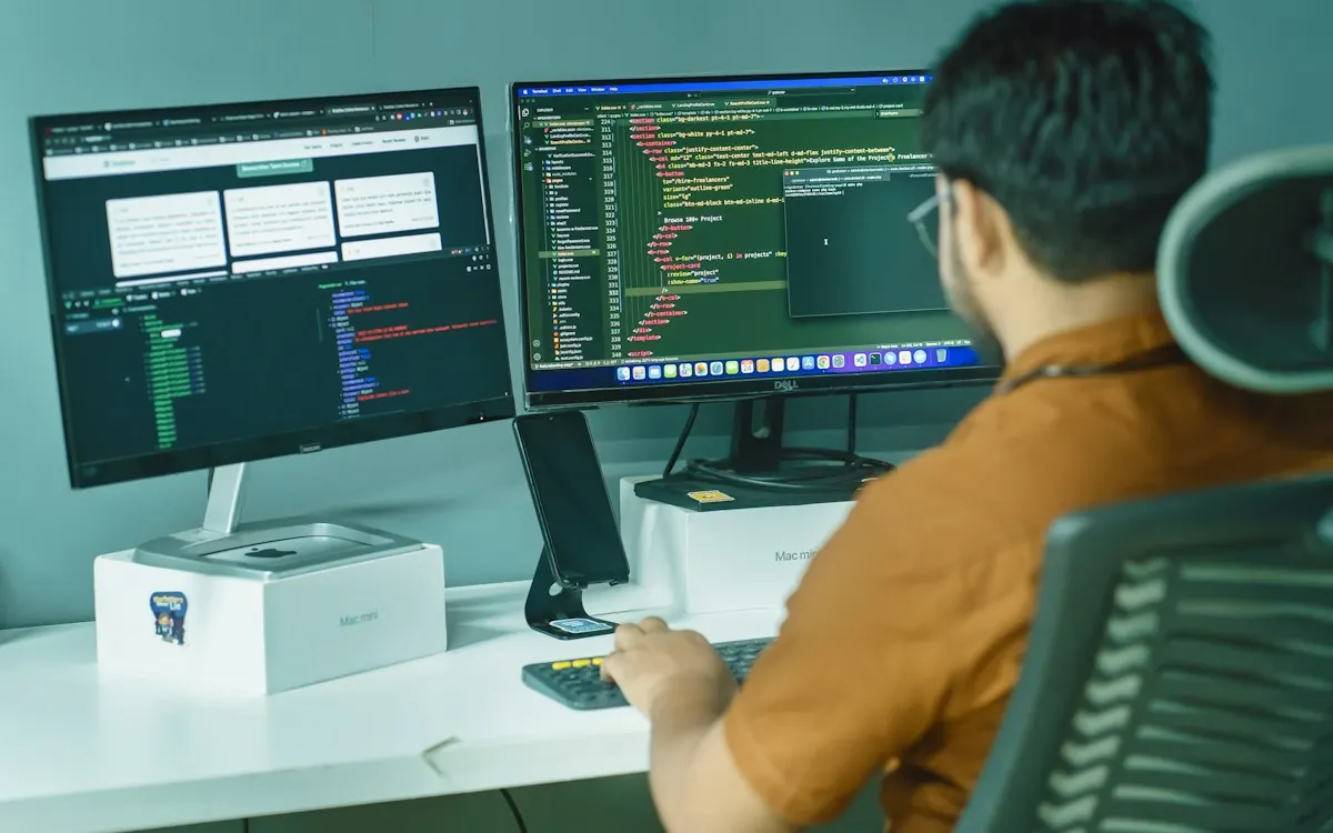 Fotografia de um homem sentado de costas numa cadeira de escritório com dois monitores de computador que exibem linhas de código e janelas de programação. O homem veste uma camisa marrom e utiliza um teclado posicionado à sua frente.